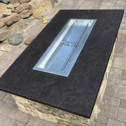 Rectangular fire pit table with a dark stone top and stainless steel drop-in burner pan installed, shown prior to adding fire glass, positioned on a paved patio with natural stone landscaping in the background.