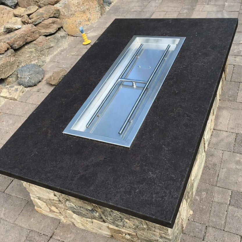 Rectangular fire pit table with a dark stone top and stainless steel drop-in burner pan installed, shown prior to adding fire glass, positioned on a paved patio with natural stone landscaping in the background.