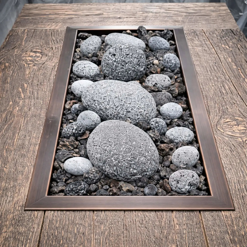 Top-down view of a rectangular drop-in burner pan framed in oil-rubbed bronze, filled with lava rock and smooth gray river stones, installed in a wood-grain fire table for a natural, rustic outdoor fire feature.