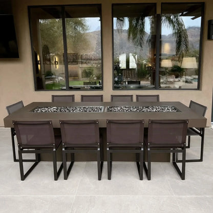 Modern outdoor dining table with an integrated rectangular fire feature, featuring two side-by-side drop-in burner pans filled with black and white fire glass, surrounded by matching patio chairs and positioned beneath a covered patio with large windows.