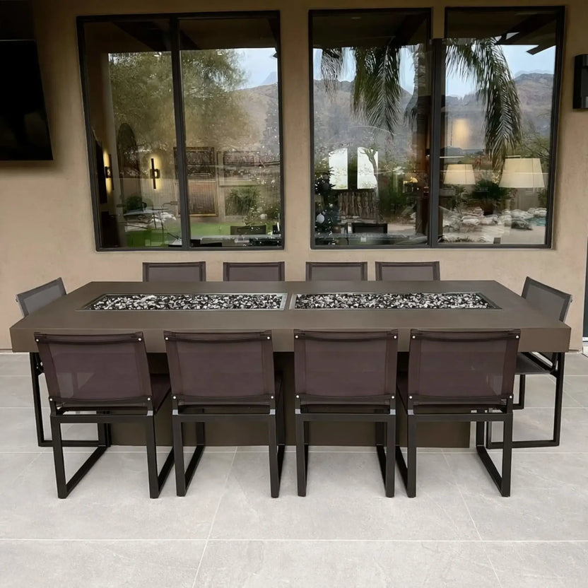 Modern outdoor dining table with an integrated rectangular fire feature, featuring two side-by-side drop-in burner pans filled with black and white fire glass, surrounded by matching patio chairs and positioned beneath a covered patio with large windows.