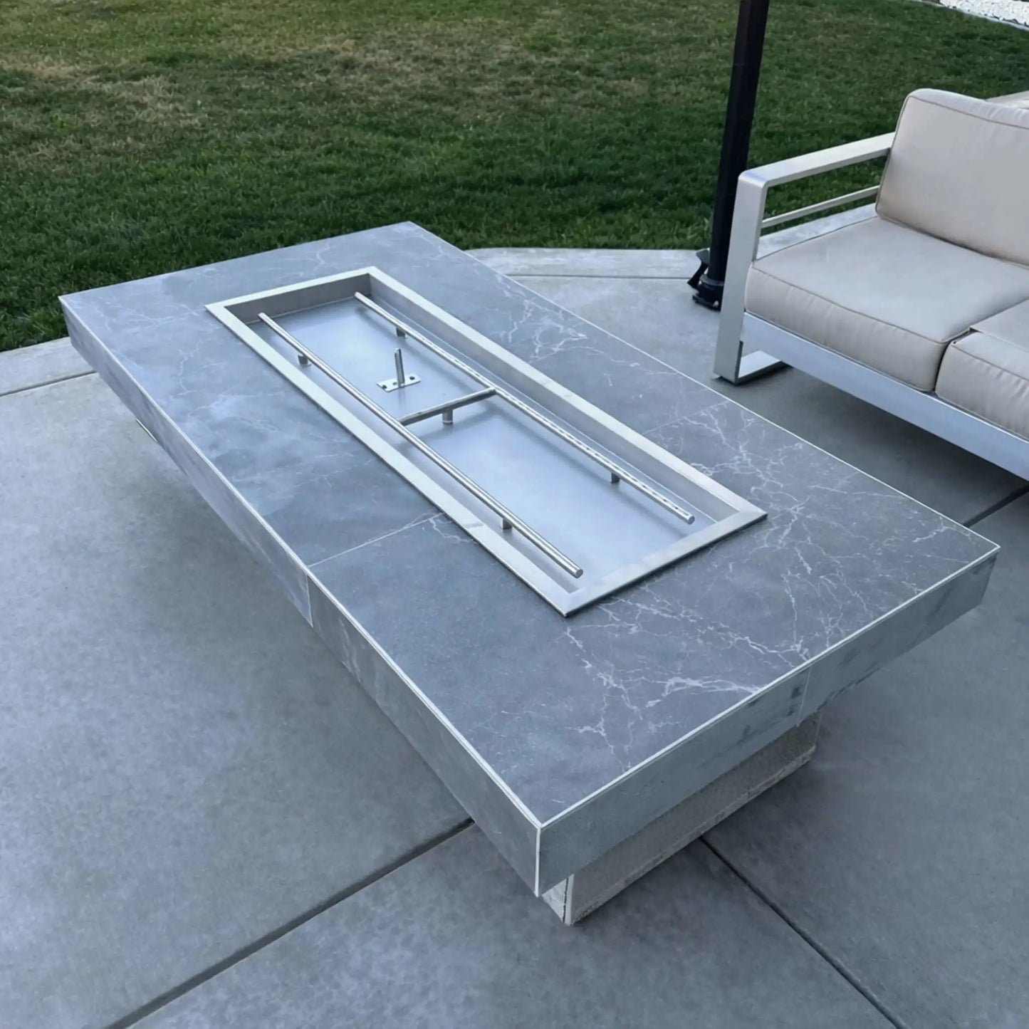 Rectangular outdoor fire pit table with a smooth gray stone finish, showcasing a stainless steel drop-in burner pan installed without fire media, set on a concrete patio next to contemporary outdoor seating and manicured lawn.