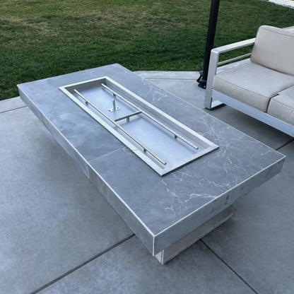 Rectangular outdoor fire pit table with a smooth gray stone finish, showcasing a stainless steel drop-in burner pan installed without fire media, set on a concrete patio next to contemporary outdoor seating and manicured lawn.