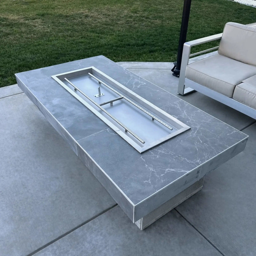 Rectangular outdoor fire pit table with a smooth gray stone finish, showcasing a stainless steel drop-in burner pan installed without fire media, set on a concrete patio next to contemporary outdoor seating and manicured lawn.