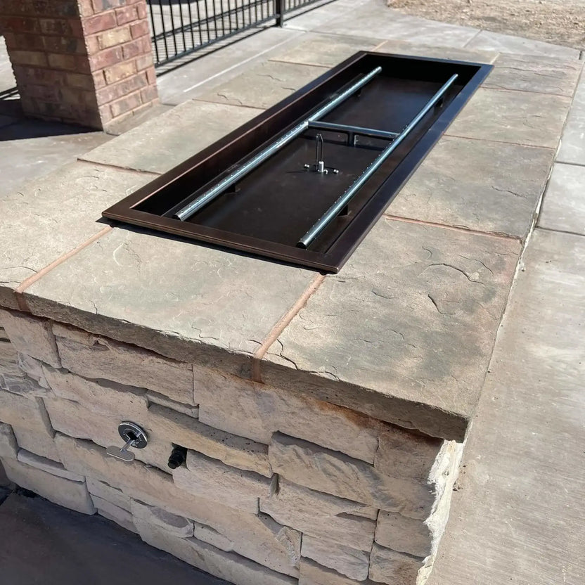 Unlit rectangular drop-in burner pan installed in a stone fire pit structure, showing the interior burner assembly and pan depth before fire glass or media is added.