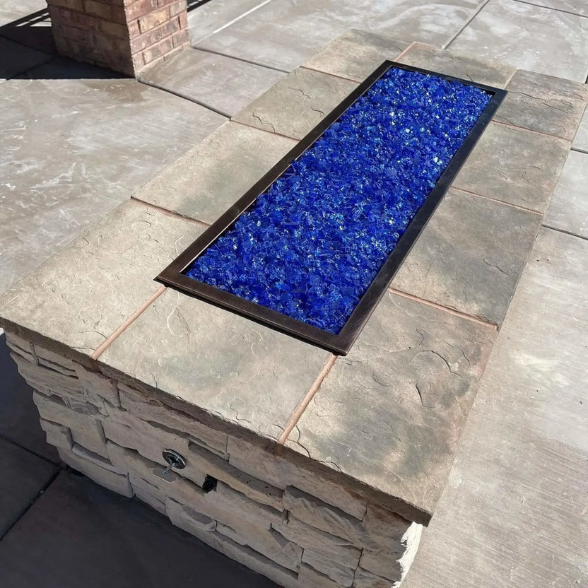 Rectangular drop-in burner pan filled with blue fire glass, installed in a stone fire pit top, showing the clean frame edges and reflective glass surface before ignition.