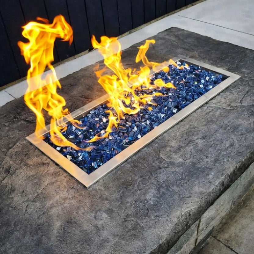 Active rectangular fire pit featuring a drop-in burner pan with blue fire glass, producing tall yellow flames against a stone patio surface in a modern outdoor living space.