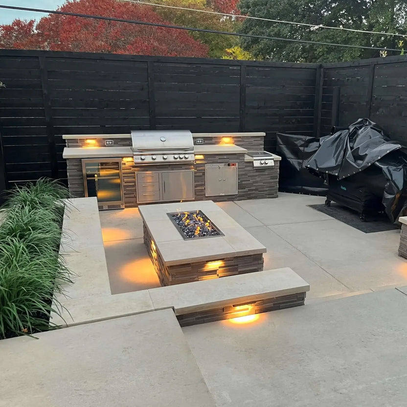 Modern backyard patio featuring a custom outdoor kitchen with stainless steel grill appliances, stone cabinetry, and a rectangular fire pit table with a drop-in burner pan centered between tiered concrete seating and integrated accent lighting.