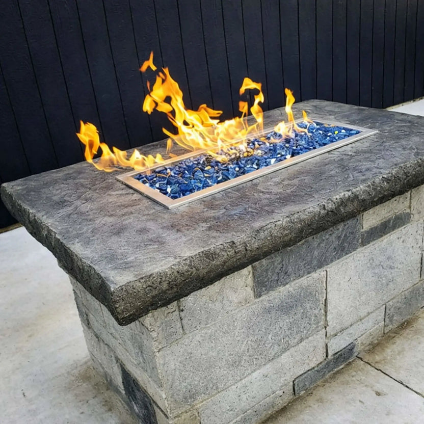 Rectangular outdoor fire pit built with stacked stone and a dark stone cap, featuring a drop-in burner pan filled with blue fire glass and active flames set against a black privacy fence.