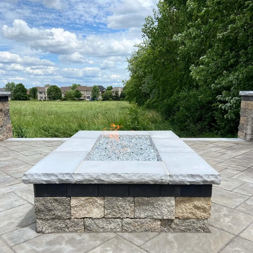 Modern outdoor fire pit featuring a rectangular flat burner pan filled with reflective fire glass, built into a stone fire pit on a paved patio with an open grassy landscape in the background.