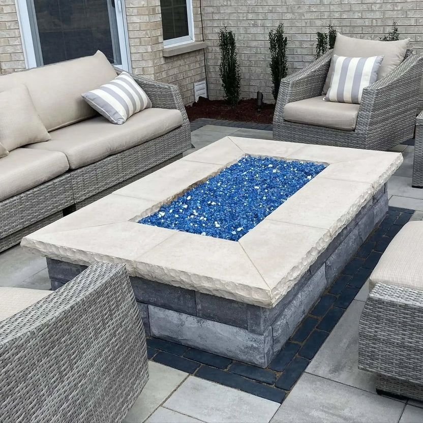 Outdoor seating area centered around a rectangular stone fire pit table using a flat stainless steel burner pan, filled with blue fire glass and surrounded by woven patio furniture and neutral stone pavers.