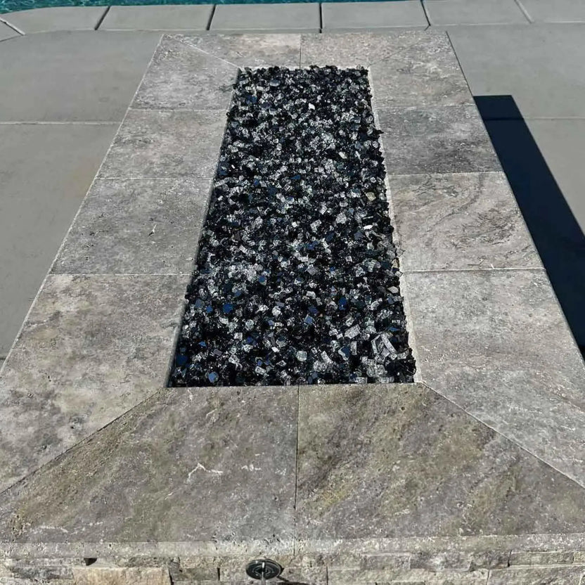 Close-up view of a rectangular fire pit burner pan filled with dark fire glass, framed by smooth stone coping and installed on a modern poolside patio with clean architectural lines.