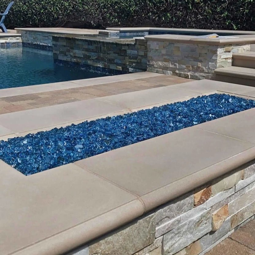 Long rectangular fire feature filled with bright blue fire glass, built into a raised stone wall beside a swimming pool, showcasing a flat stainless steel burner pan in a high-end outdoor living space.