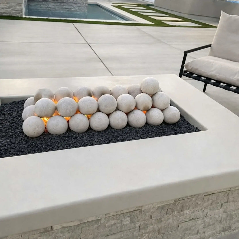 Modern rectangular fire pit table featuring a flat burner pan with decorative fire spheres over black lava rock, set within a sleek outdoor patio design with minimalist seating and light stone finishes.