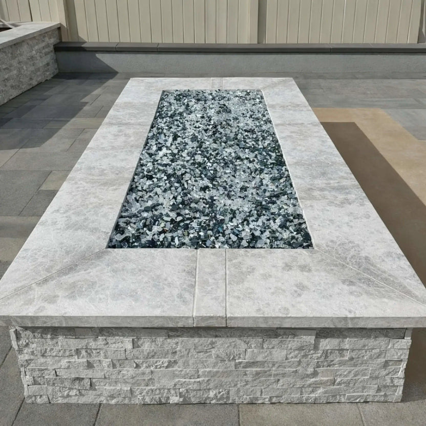 Rectangular outdoor fire pit with a light stone cap and stacked stone base, featuring a recessed stainless steel flat burner pan filled with gray fire glass. The fire pit sits on a paved patio in a clean, modern backyard setting with neutral tones and minimalist landscaping.