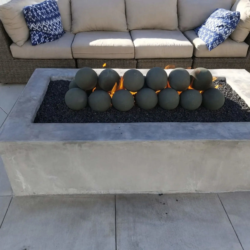 Contemporary rectangular fire pit with a smooth concrete exterior and a stainless steel flat burner pan filled with black lava rock. Decorative ceramic fire balls are stacked over the burner, with visible flames creating a warm focal point in front of an outdoor sectional sofa.