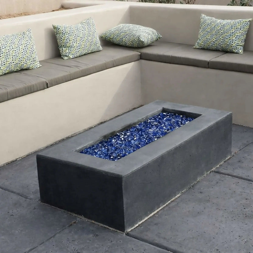 Modern concrete fire pit featuring a long rectangular stainless steel burner pan filled with blue fire glass. The fire pit is positioned in a sunken outdoor seating area with built-in benches, gray cushions, and patterned accent pillows for a clean, contemporary look.