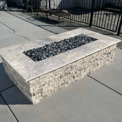Rectangular stone fire pit with a light natural stone cap and textured stacked stone sides, showcasing a stainless steel flat burner pan filled with dark fire glass. The fire pit is installed beside a pool deck with concrete pavers and black metal fencing in the background.