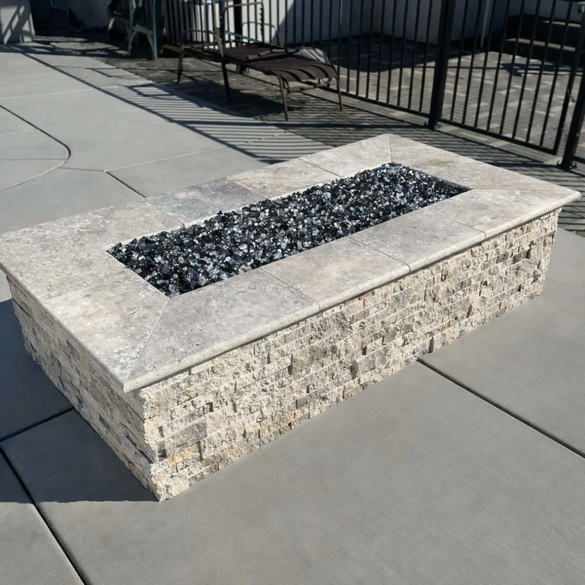 Rectangular stone fire pit with a light natural stone cap and textured stacked stone sides, showcasing a stainless steel flat burner pan filled with dark fire glass. The fire pit is installed beside a pool deck with concrete pavers and black metal fencing in the background.