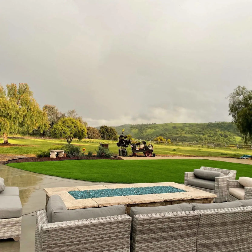 Rectangular stone fire pit with a stainless steel flat burner pan and blue fire glass, positioned in a luxury backyard setting with pool water, palm trees, and sunset lighting.