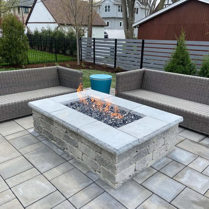 Outdoor patio seating area with a rectangular fire pit table using a flat stainless steel burner pan, filled with smooth lava stones and producing a clean, linear flame.