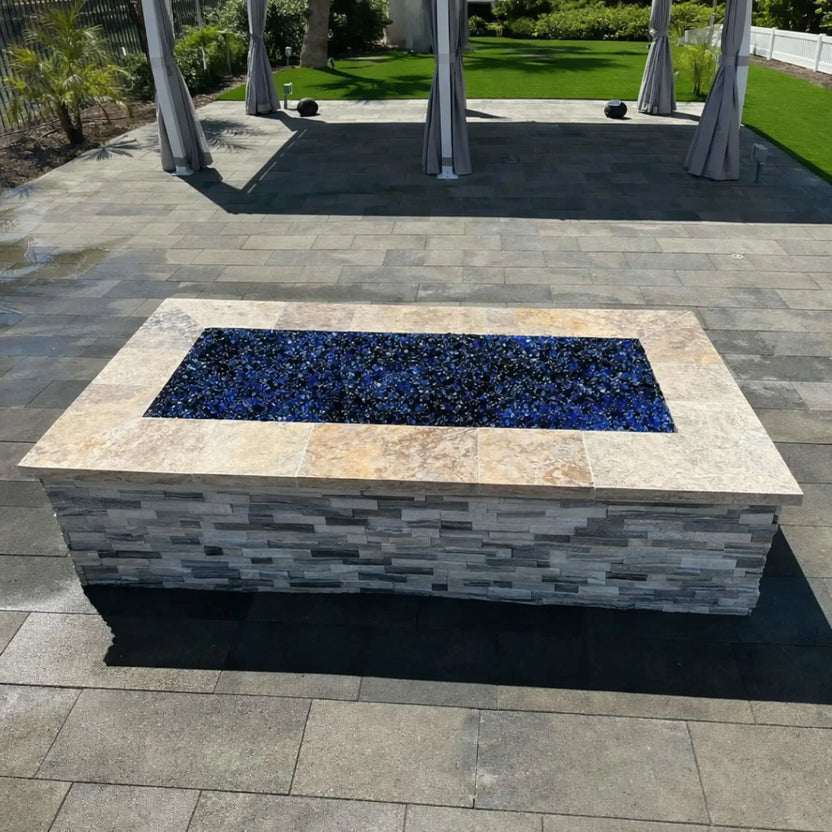 Rectangular stone fire pit with a flat stainless steel burner pan filled with deep blue fire glass, installed on a large paved patio beneath a shaded pergola, surrounded by manicured lawn and modern outdoor landscaping.