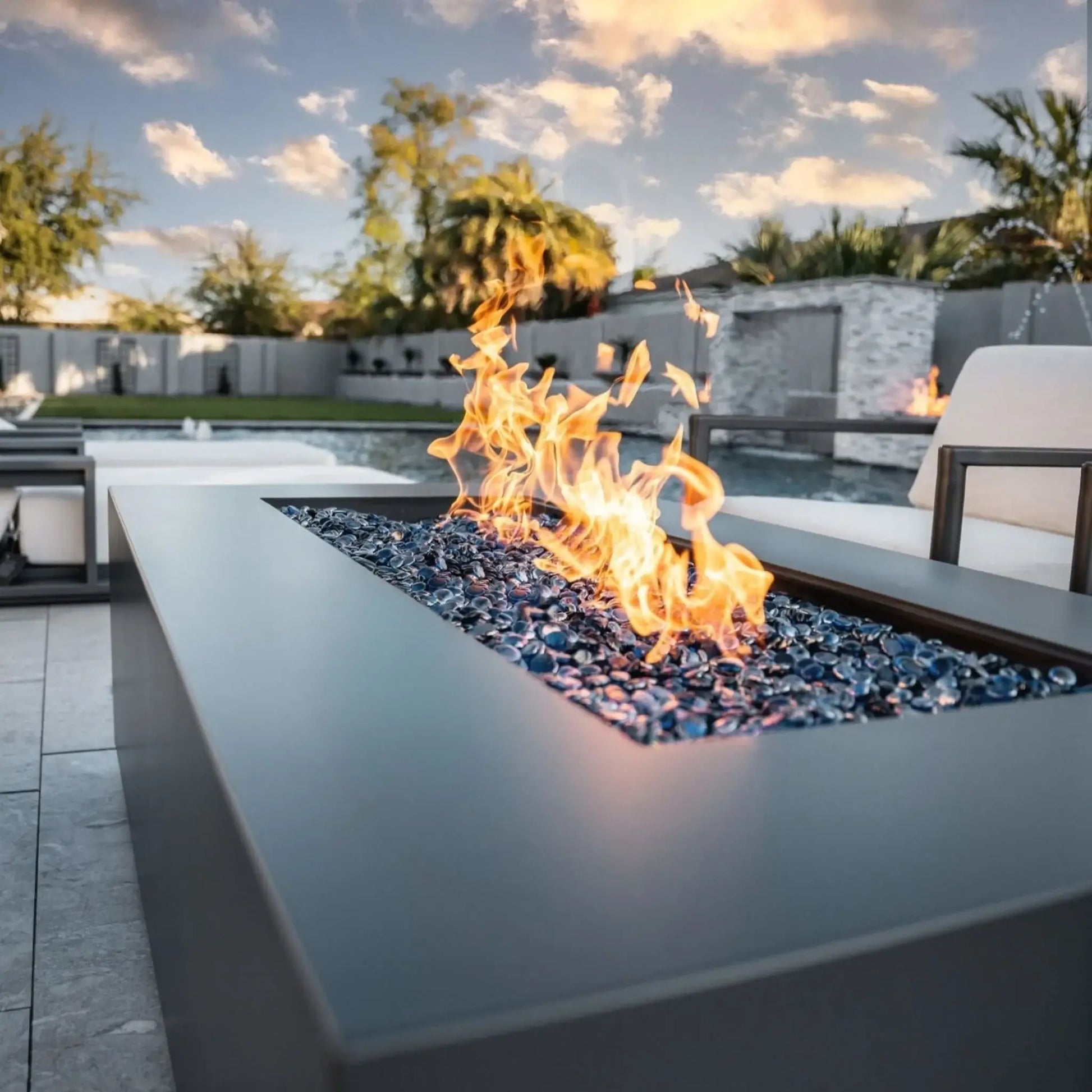 Contemporary rectangular fire pit table featuring a flat stainless steel burner pan with blue fire glass and active flames, positioned beside a pool in a luxury backyard with lounge seating and palm trees at sunset.