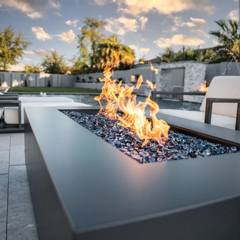 Contemporary rectangular fire pit table featuring a flat stainless steel burner pan with blue fire glass and active flames, positioned beside a pool in a luxury backyard with lounge seating and palm trees at sunset.
