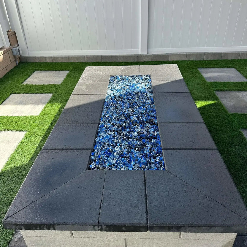 Rectangular fire pit built with smooth stone coping and a flat burner pan filled with blue reflective fire glass, set into a clean, modern patio design with white fencing and artificial turf accents.