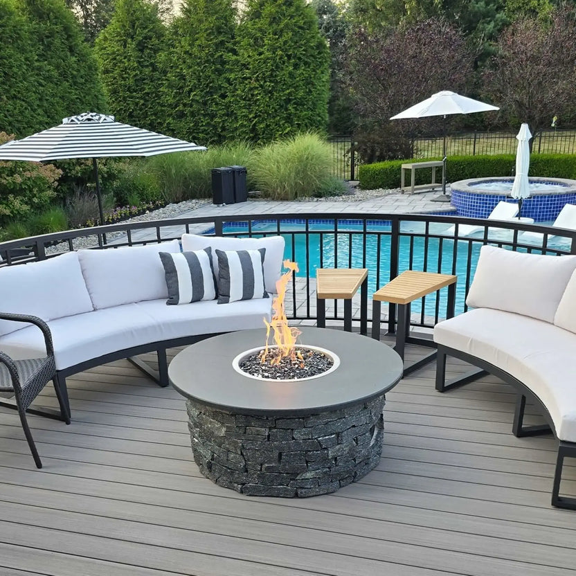 Round stainless steel drop-in fire pit burner pan installed in a stone fire pit table on a poolside deck, featuring a bright natural gas flame over dark fire glass with curved outdoor seating and a swimming pool in the background.