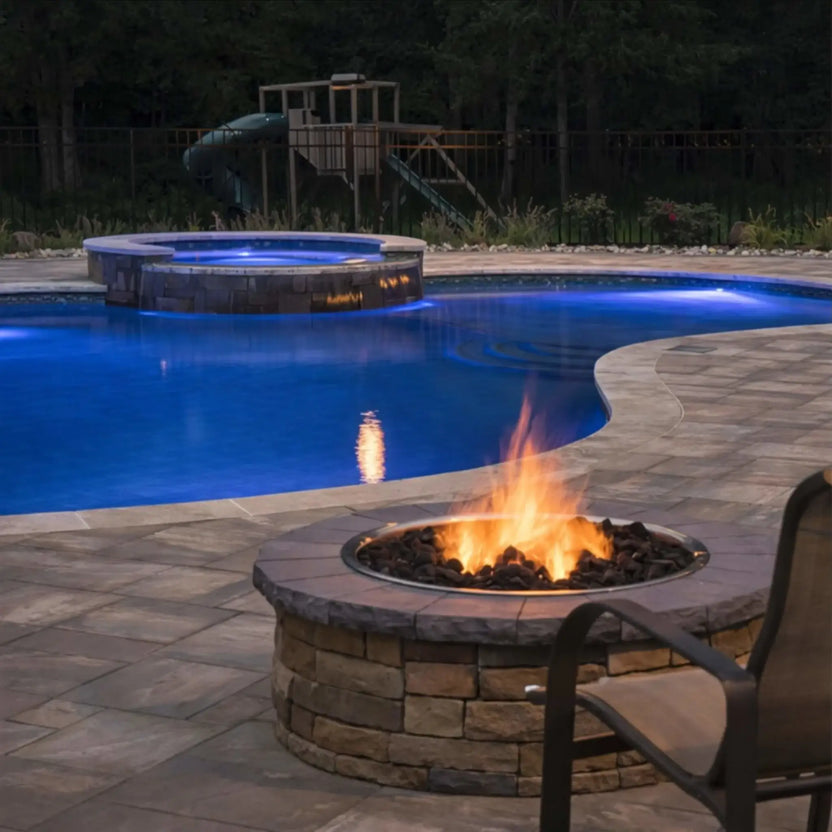 Circular stainless steel fire pit burner pan burning with a steady flame beside a luxury backyard swimming pool at night, showcasing stone masonry construction, fire glass media, and soft ambient pool lighting.