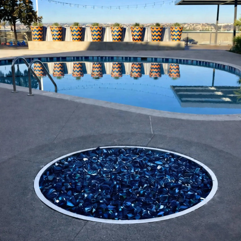 Round drop-in fire pit burner pan filled with blue fire glass, installed flush into a concrete patio near a modern swimming pool, highlighting a clean stainless steel trim and contemporary outdoor design.