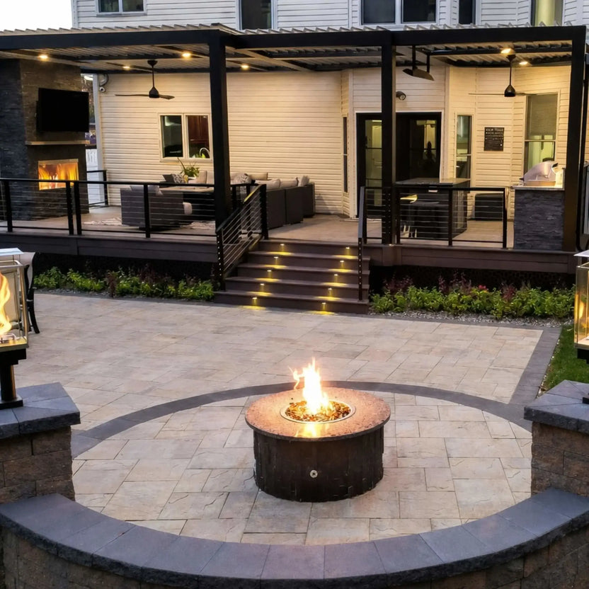 Modern backyard patio with a round stainless steel fire pit burner pan set into stone pavers, positioned in front of a covered deck with integrated lighting, outdoor seating, and a contemporary pergola structure.