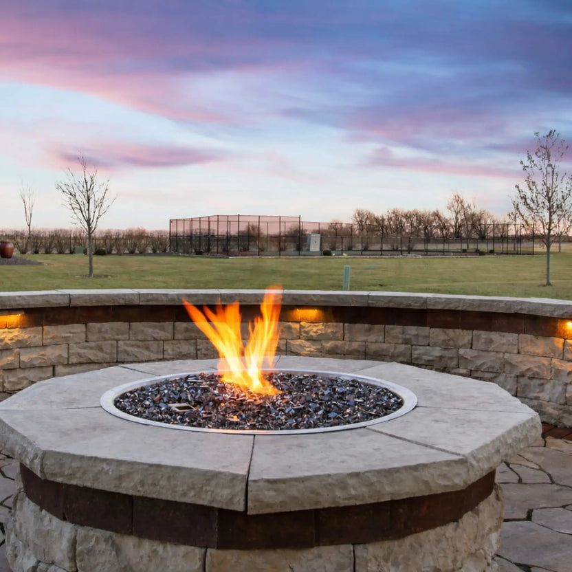 Close-up of a round stone fire pit featuring a stainless steel drop-in burner pan with active flames over fire glass, set against an open landscape at sunset with a curved retaining wall and outdoor accent lighting.