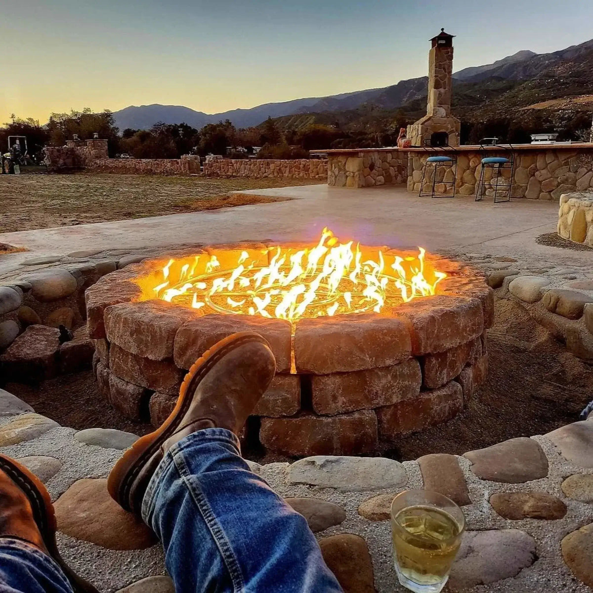 Outdoor stone fire pit with a stainless steel round burner ring producing bright yellow flames at sunset, viewed from a relaxed seated perspective with boots in the foreground, mountain landscape and outdoor kitchen patio in the background.