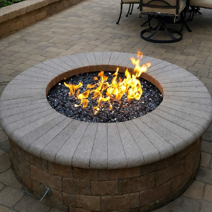 Outdoor round fire pit constructed with stone block seating wall and a stainless steel flat burner pan filled with dark fire glass, producing vibrant flames in a paved patio setting with curved masonry bench seating.