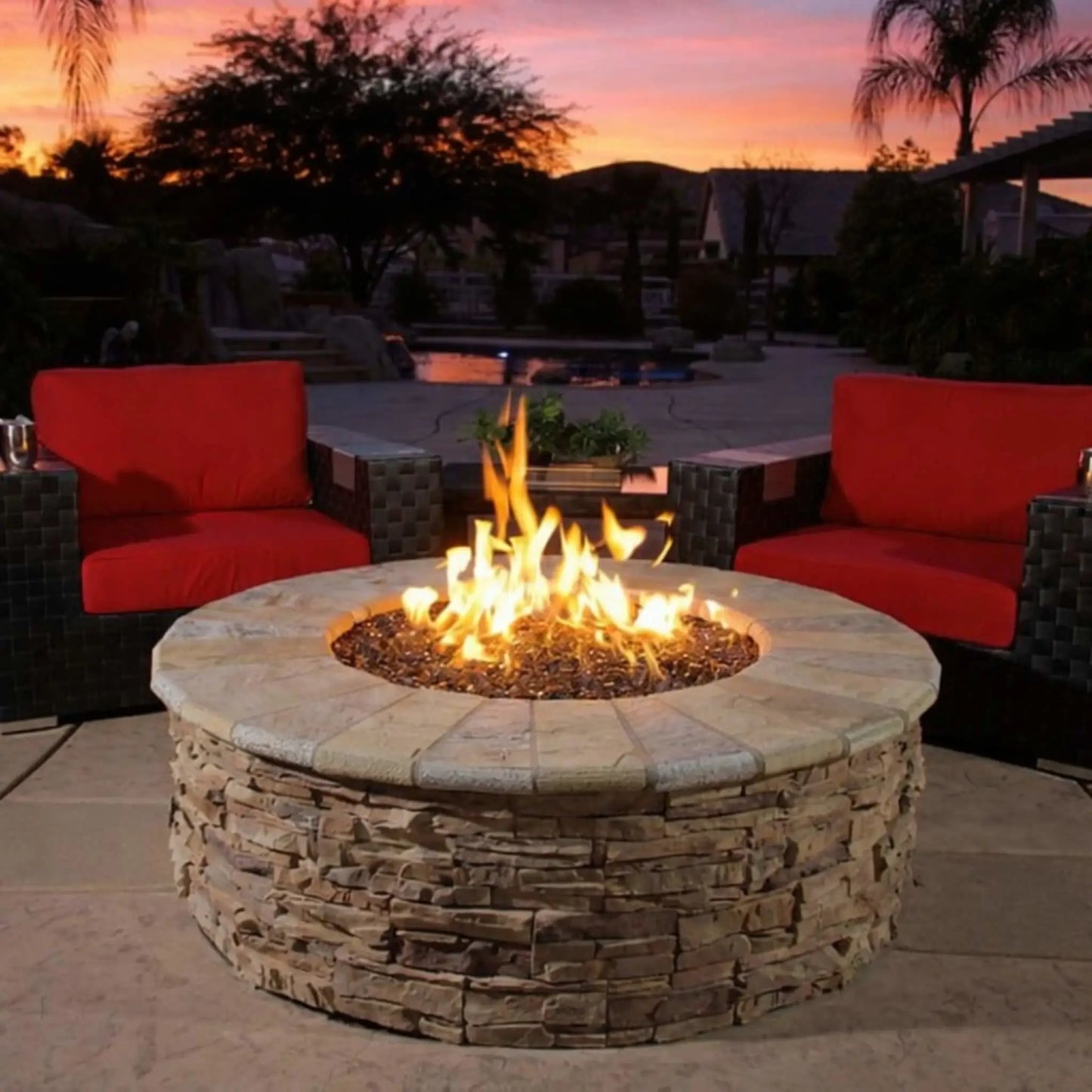 Round stone fire pit with bright natural gas flames rising from fire glass, centered between modern outdoor lounge chairs on a luxury patio at sunset, creating a warm and inviting backyard entertaining space.