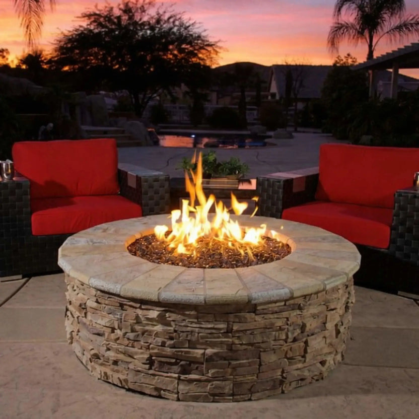 Round stone fire pit with bright natural gas flames rising from fire glass, centered between modern outdoor lounge chairs on a luxury patio at sunset, creating a warm and inviting backyard entertaining space.