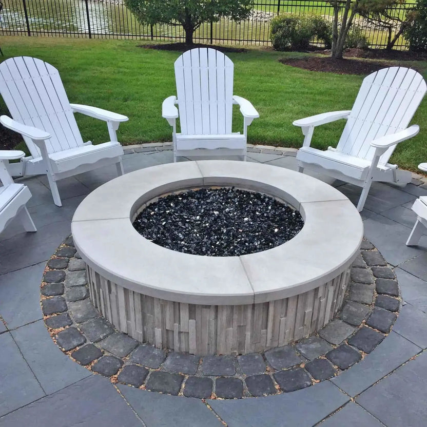 Close-up nighttime view of a round outdoor fire pit featuring reflective fire glass and tall dancing flames, set into a textured stone fire pit surround for dramatic evening ambiance.