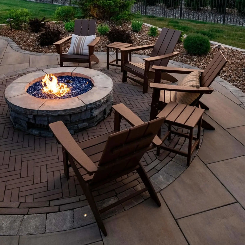 Round masonry fire pit filled with black fire glass, surrounded by white Adirondack chairs on a circular paver patio, showcasing a clean and modern backyard fire pit design during daylight.