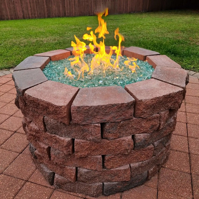 Round masonry fire pit built with stacked stone blocks, filled with aqua fire glass and active gas flames, set on a brick paver patio with green lawn and wooden privacy fence in the background.