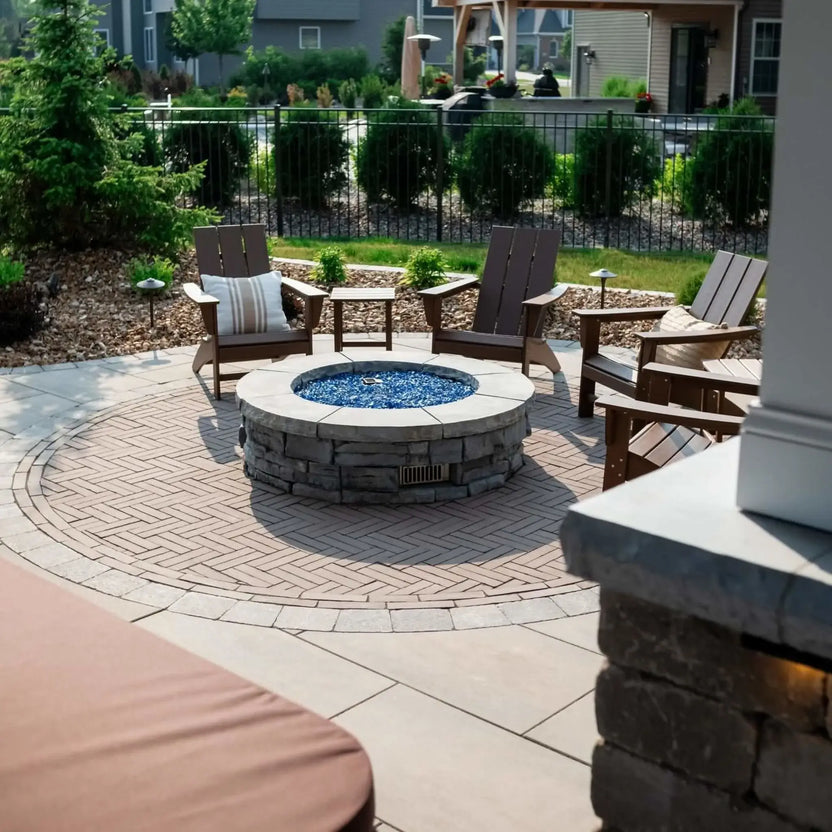 Outdoor circular gas fire pit featuring blue fire glass media, surrounded by Adirondack chairs on a patterned paver patio in a landscaped residential courtyard setting.