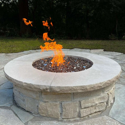 Modern round stone fire pit with a smooth concrete cap and reflective fire glass, showing tall orange flames rising at the center on a natural stone patio surrounded by trees.