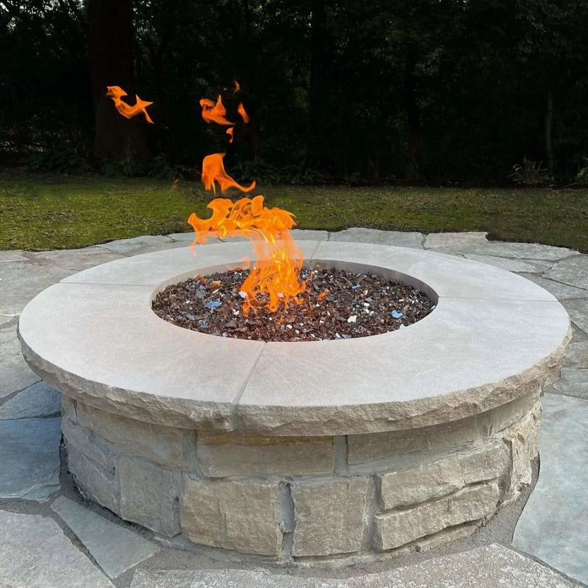 Modern round stone fire pit with a smooth concrete cap and reflective fire glass, showing tall orange flames rising at the center on a natural stone patio surrounded by trees.