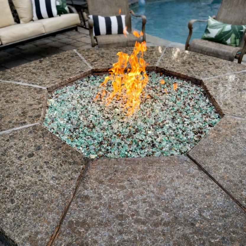 Close-up view of a luxury outdoor fire pit table with polished stone surface and green fire glass, producing vibrant gas flames beside a pool and cushioned patio seating.