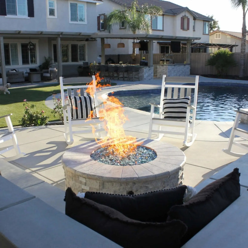 Round stone fire pit with a stainless steel burner pan filled with blue fire glass, producing tall natural gas flames on a modern backyard patio with white rocking chairs and a swimming pool in the background.