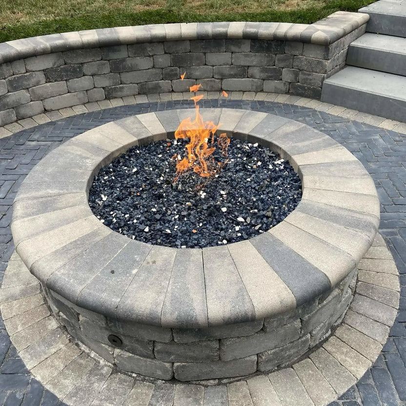 Circular stone fire pit featuring a stainless steel flat burner pan beneath black fire glass media, showcasing steady gas flames centered within a landscaped hardscape patio and curved retaining wall design.