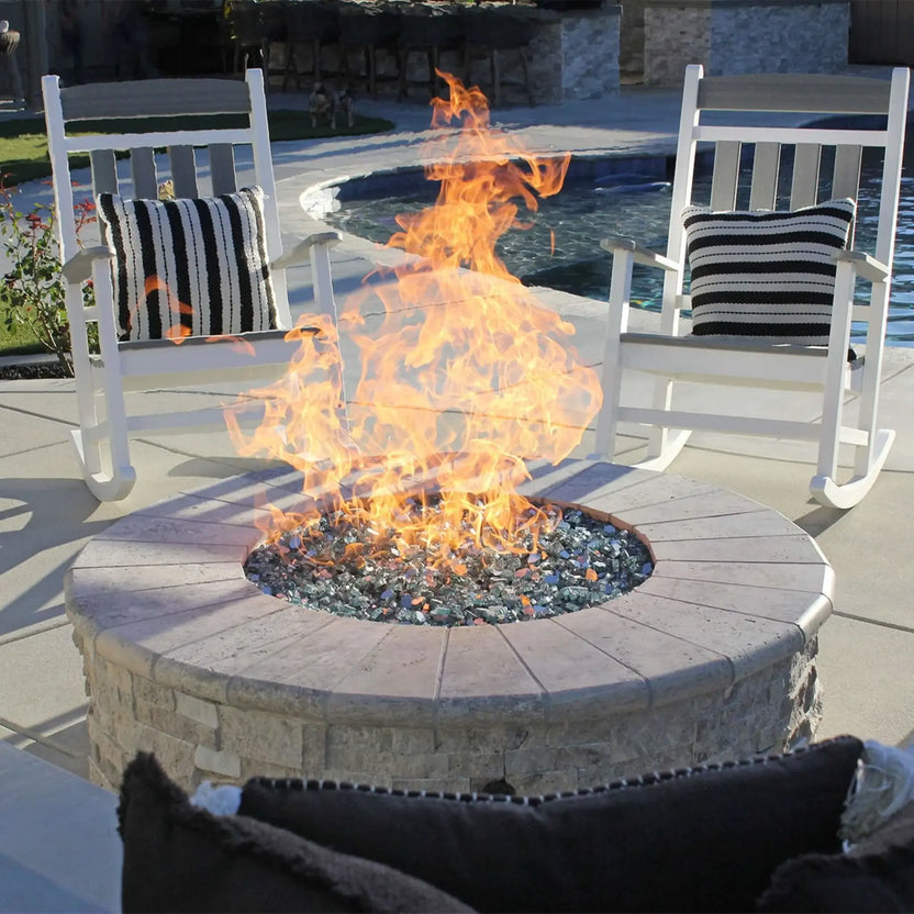 Close-up view of a round masonry fire pit featuring a stainless steel flat burner pan and reflective fire glass, with bright flames rising between two white outdoor rocking chairs beside a residential pool.