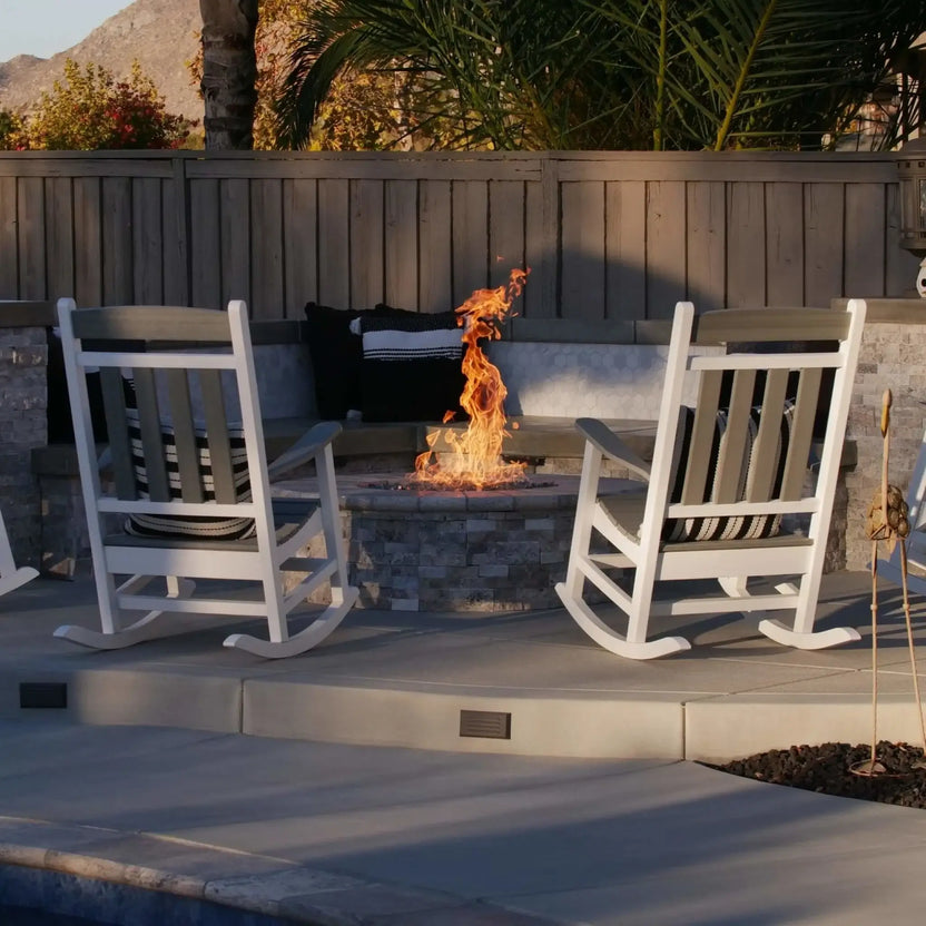 Outdoor seating area with a circular stone fire pit centered between white rocking chairs, showcasing a stainless steel burner pan with fire glass and tall flames against a privacy fence and landscaped backyard.