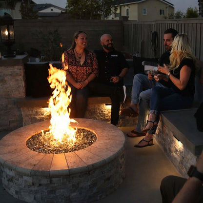 Evening backyard gathering around a round stone fire pit with a stainless steel burner pan and fire glass, where friends relax on built-in seating as warm flames illuminate the outdoor living space.